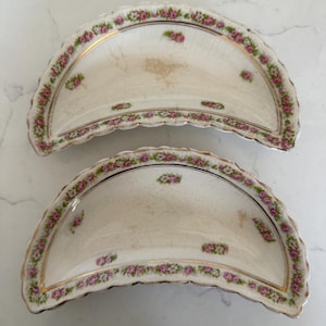 May include: Two crescent-shaped ceramic bone dishes with a cream-colored base, featuring a scalloped edge and a floral pattern of pink roses and green leaves. The dishes have a gold-trimmed border and visible surface crazing throughout.