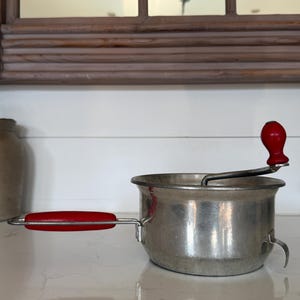 May include: A silver-toned metal food mill featuring a cylindrical basin, a side handle with a red grip, and a top-mounted hand crank with a matching red knob. The device includes a small metal hook on the side for securing it to a bowl or pot.