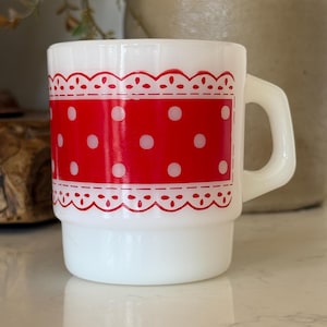 May include: A white milk glass mug featuring a wide horizontal band of bright red with a repeating pattern of white polka dots. The red band is bordered on the top and bottom by a decorative red scalloped lace-like trim design.
