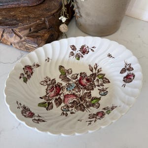 May include: An oval-shaped white ceramic bowl featuring a scalloped, fluted edge and a central floral pattern. The design includes detailed illustrations of blooming roses in shades of pink and deep red, surrounded by brown leaves and stems.