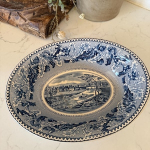 May include: An oval ceramic bowl featuring a blue and white transferware pattern. The center depicts a landscape with a canal, boat, and buildings. The rim is decorated with a detailed border of oak leaves and acorns on a mesh background.