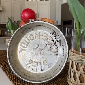 May include: A vintage, round, metal pie pan with embossed text and a star design in the center. The pan has a distressed, aged appearance with signs of wear and rust. The text reads "YOOM-E 2719".