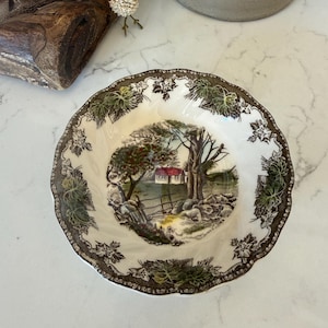 May include: A round ceramic bowl with a scalloped edge featuring a central landscape illustration of a house with a red roof, trees, and a stone wall. The rim is decorated with a dark brown and green grape leaf pattern on a cream background.
