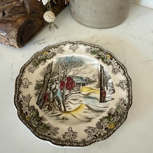 May include: A round ceramic plate with a scalloped edge featuring a brown and white floral border. The center depicts a winter landscape with a red barn, bare trees, and buckets attached to tree trunks, rendered in muted tones of red, blue, and yellow.