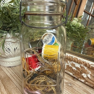 May include: A clear glass jar with a metal handle and lid, filled with sewing supplies. The jar contains spools of thread in various colors, gold-colored trim, and pink ribbon. The jar is sitting on a wooden surface.