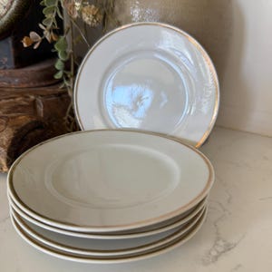 May include: A stack of four round white ceramic saucers, each featuring a thin metallic gold rim along the edge. The plates have a smooth, glossy finish and a slightly recessed center, presented in a clean, uniform arrangement for dining use.