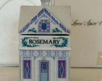 May include: A white porcelain spice jar shaped like a house with a roof lid. The front features the word ROSEMARY in black text, surrounded by green vines. The house has blue and purple accents on the windows, door, and roof trim details.