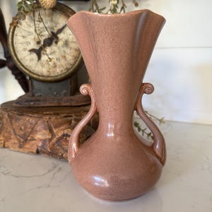 May include: A vintage, speckled, pink vase with a flared top and two curved handles. The vase has a rounded base and a textured surface. The design is reminiscent of mid-century modern decor. The vase is likely made of ceramic or pottery.