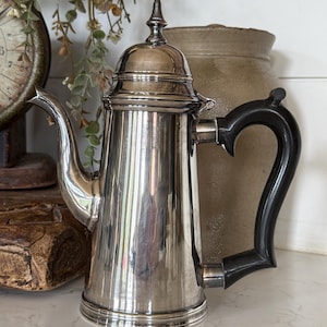 May include: A silver-toned metal coffee pot featuring a tall, cylindrical body, a curved spout, and a black handle. The pot includes a matching lid with a pointed finial top. The surface of the metal is reflective and smooth, showing vertical light reflections.