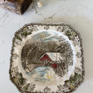 May include: A square ceramic plate with a scalloped edge featuring a brown and white floral border. The center depicts a winter landscape with a red-roofed covered bridge over a stream, surrounded by snow-covered trees and a mountain background.