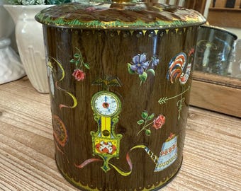 May include: A vintage, round, metal tin with a gold lid and a woodgrain design. The tin is decorated with colorful floral and geometric patterns, including a clock, a rooster, and a teapot.