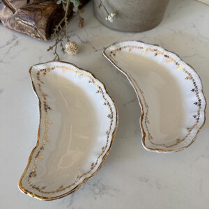 May include: Two crescent-shaped white porcelain dishes featuring scalloped edges and delicate gold floral detailing along the rims. The dishes have a smooth, glossy finish and a curved, shell-like form, showcasing intricate gold leaf patterns.