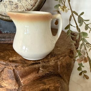 May include: A small, off-white ceramic creamer with a tan rim and a handle. The creamer sits on a carved wooden surface, with a vintage scale in the background. Decorative greenery hangs on the right side.