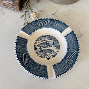 May include: A round ceramic ashtray featuring a white center with a blue transferware illustration of two figures on a bench in a park. The wide blue rim is decorated with hatched lines, scrollwork, and a beaded edge, including three notches.
