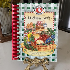 May include: A red and green spiral bound cookbook with a green plaid cover. The cover features a goose illustration and the text "Gooseberry Patch's Christmas Pantry". The book is on a gold metal stand.
