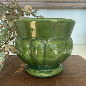 May include: A glossy, green ceramic planter with a scalloped design around the body. The planter has a wide opening and sits on a small, round base. The surface reflects light, highlighting its curves. A decorative piece for indoor plants.