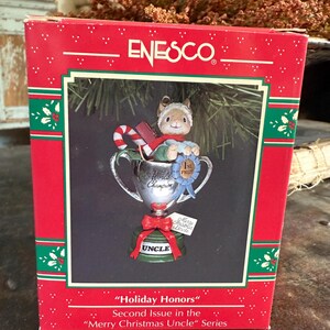 May include: A red and green box featuring an Enesco Christmas ornament. The ornament depicts a small mouse in a trophy with a candy cane. The trophy has a blue ribbon that says "1st Prize". The box reads "Holiday Honors" and "Merry Christmas Uncle".