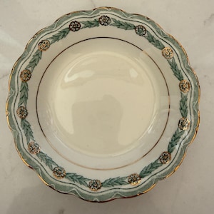 May include: A cream-colored, scalloped-edge plate with a green and gold floral border. The border features a repeating pattern of green leaves and gold flowers. The rim is trimmed with a thin gold band.