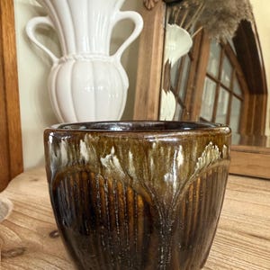 May include: A brown and white glazed ceramic planter with vertical grooves. The planter has a glossy finish and a rounded shape. A white vase with handles is in the background.