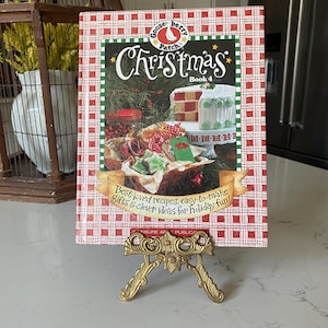 May include: A red and white checkered cookbook titled "Gooseberry Patch Christmas Book 4" with a white goose logo and the text "Best-loved recipes, easy-to-make gifts & clever ideas for holiday fun!" The book is displayed on a gold metal stand.