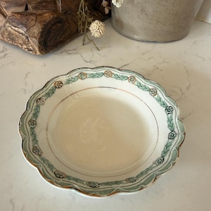 May include: A shallow, round ceramic bowl with a scalloped edge. The rim features a decorative pattern of green leafy garlands interspersed with small, circular floral motifs in shades of brown and blue, accented by a thin gold-toned trim.