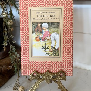May include: A vintage book titled "The Fir Tree" by Hans Christian Andersen, illustrated by Nancy Ekholm Burkert. The book has a red patterned cover and is displayed on a decorative gold-colored stand. The book's illustration depicts children in a forest.