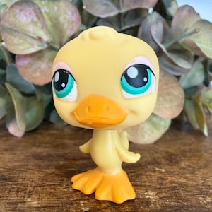 May include: A yellow plastic duck figurine with large, expressive eyes. The duck has an orange beak and feet, and its body is a lighter shade of yellow. The figurine is standing on a wooden surface, with greenery in the background.