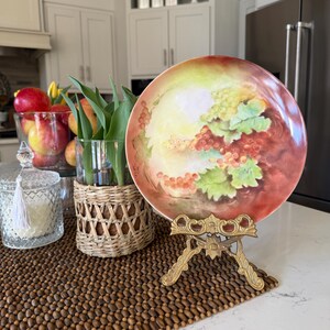 May include: A decorative plate with a hand-painted grape design, displayed on a gold-colored metal stand. The plate features red, green, and yellow hues. The plate is set on a brown beaded mat, with a glass vase of tulips and a glass jar with a tassel.