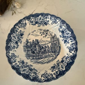 May include: A round ceramic bowl with a scalloped edge featuring a blue and white transferware pattern. The center depicts a horse-drawn stagecoach scene, while the border is decorated with intricate floral motifs and scrollwork.
