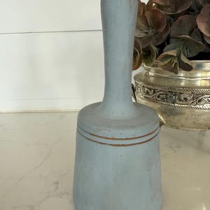 May include: A light blue, wooden kitchen muddler with a rounded base and a long handle. The muddler has a distressed finish with visible wood grain and a touch of gold-colored accents. The kitchen tool is designed for crushing herbs and fruits.