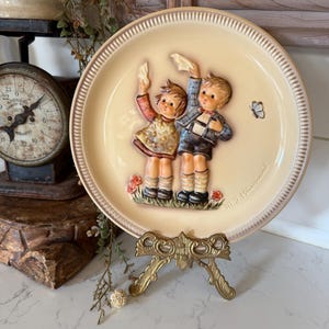 May include: A decorative ceramic plate with a cream-colored background and a raised relief design. The plate features two children, a boy and a girl, holding up handkerchiefs. A small butterfly and flowers are also depicted.
