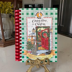 May include: A cookbook titled "Coming Home for Christmas" with a green plaid cover and red spiral binding. The cover features a festive illustration of a decorated doorway. The book is displayed on a gold-colored easel.