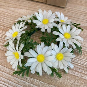 May include: A white daisy flower wreath with green leaves. The wreath is made of artificial flowers and is perfect for decorating a home or adding a touch of nature to a space.