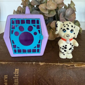 May include: A vintage toy set featuring a dalmatian puppy figurine and a purple and teal pet carrier. The puppy has black spots, blue eyes, and a red collar. The carrier has a paw print design on the door.