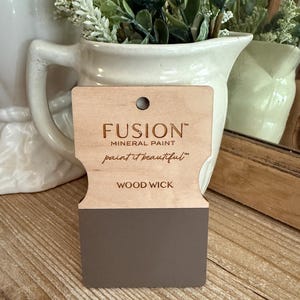May include: A wooden paint swatch from Fusion Mineral Paint, labeled "Wood Wick." The swatch displays a muted, brownish-gray color. The brand name and slogan "paint it beautiful" are printed on the top portion of the swatch.