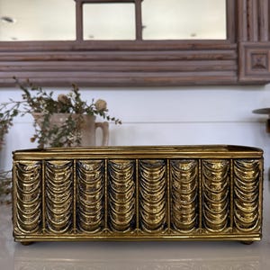 May include: A rectangular, gold-colored decorative box with a detailed, textured design. The box features a series of vertical, curved patterns along its sides, giving it a vintage or antique appearance. The box is elevated on small feet.