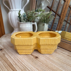 May include: A double-sectioned, yellow ceramic planter with a woven basket design. The planter is a bright, sunny yellow and sits on a wooden surface. Other white ceramic vases and a mirror are in the background.