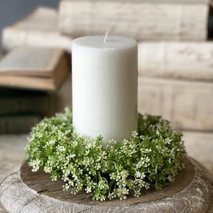May include: A white pillar candle sits atop a wooden base, encircled by a wreath of small green and white flowers. The candle is unlit, and the base has a rustic, weathered appearance. The background features stacks of old books.