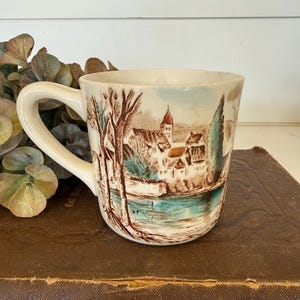 May include: Cream-colored ceramic mug featuring a scenic village landscape with a river, trees, and buildings. The handle is curved, and the mug sits on a brown surface. The artwork includes shades of brown, blue, and green.