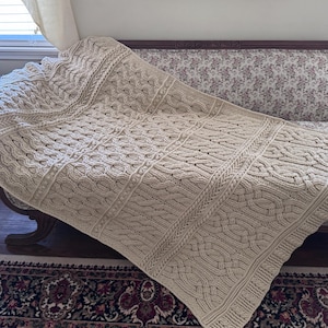 May include: A beige, hand-knitted blanket with intricate cable and textured stitch patterns. The blanket is draped over a floral-patterned sofa, showcasing its detailed craftsmanship and cozy aesthetic. The blanket's dimensions are approximately 60 inches by 80 inches.