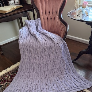 May include: A lavender-colored knitted blanket with a cable knit design draped over a vintage pink upholstered chair. The blanket's texture and pattern are visible, creating a cozy and inviting aesthetic. The blanket is resting on a patterned rug.