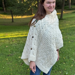 May include: An off-white, cable-knit poncho featuring a textured basketweave pattern and a high collar. The garment is secured along the side with three round, dark brown buttons, creating a structured drape over the shoulders and torso.
