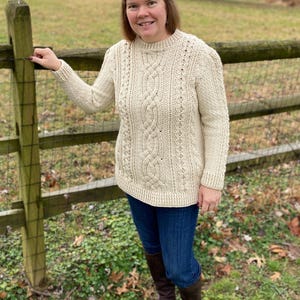 Può includere: Una donna indossa un maglione di lana a treccia color crema con scollo rotondo. È in piedi davanti a una recinzione di legno e sorride alla telecamera.