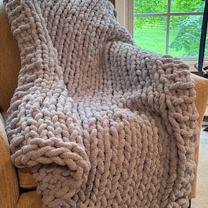 May include: A chunky knit blanket in a soft gray color is draped over a beige armchair. The blanket's thick, looped texture is visible, creating a cozy and inviting appearance. The background includes a window with a view of greenery.