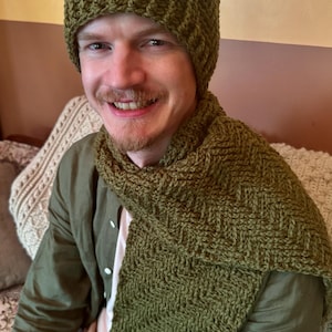 May include: A matching olive green set consisting of a knit beanie and a long scarf. Both items feature a textured, repeating chevron or herringbone pattern created with thick yarn. The accessories are uniform in color and stitch design.