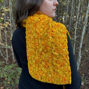 May include: A yellow and orange knitted scarf with a cable knit pattern. The scarf is worn by a person in a forest setting.