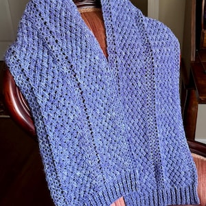 May include: A blue knitted shawl with a V-neck design. The shawl is made of a soft, textured yarn and is draped over a chair. The knitted pattern is visible throughout the shawl.