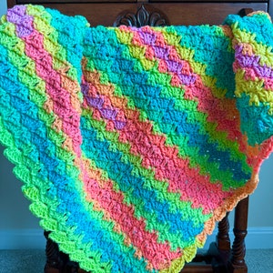 Op de afbeelding: Een kleurrijk gehaakt babydekentje met een gegolfde rand. Het dekentje is gemaakt van verschillende felle kleuren, waaronder roze, blauw, groen en geel.