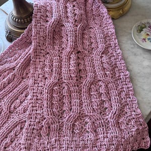May include: A dusky pink knitted scarf featuring a detailed cable and basketweave pattern. The scarf is draped, highlighting its intricate texture and design. The handmade scarf is a cosy accessory, perfect for cooler weather.