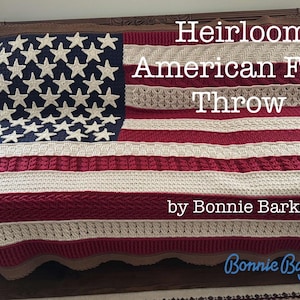 Op de afbeelding: Een gehaakte plaid met het ontwerp van de Amerikaanse vlag in rood, wit en blauw garen. De tekst op de deken luidt: Heirloom American Flag Throw by Bonnie Barker, Bonnie Bay Crochet. De deken heeft een gestructureerd kabelpatroon.